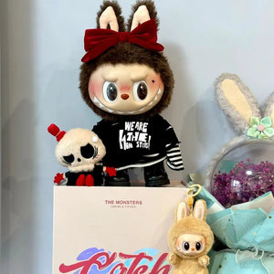 Plush toy with bunny ears and a black shirt, standing next to a box with colorful text, surrounded by other plush toys.